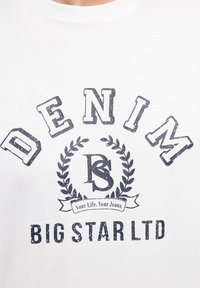 Balts kokvilnas T-krekls ar tumši zilu vintage stila tekstu, kurā ir rakstīts "DENIM", lauru dizains un zem tā "BIG STAR LTD".