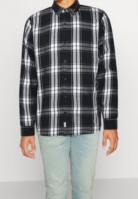 Svart och vit rutig skjorta med grå inslag, knappad krage, lång ärm och en sydd etikett nära fållen, matchad med ljusblå jeans.