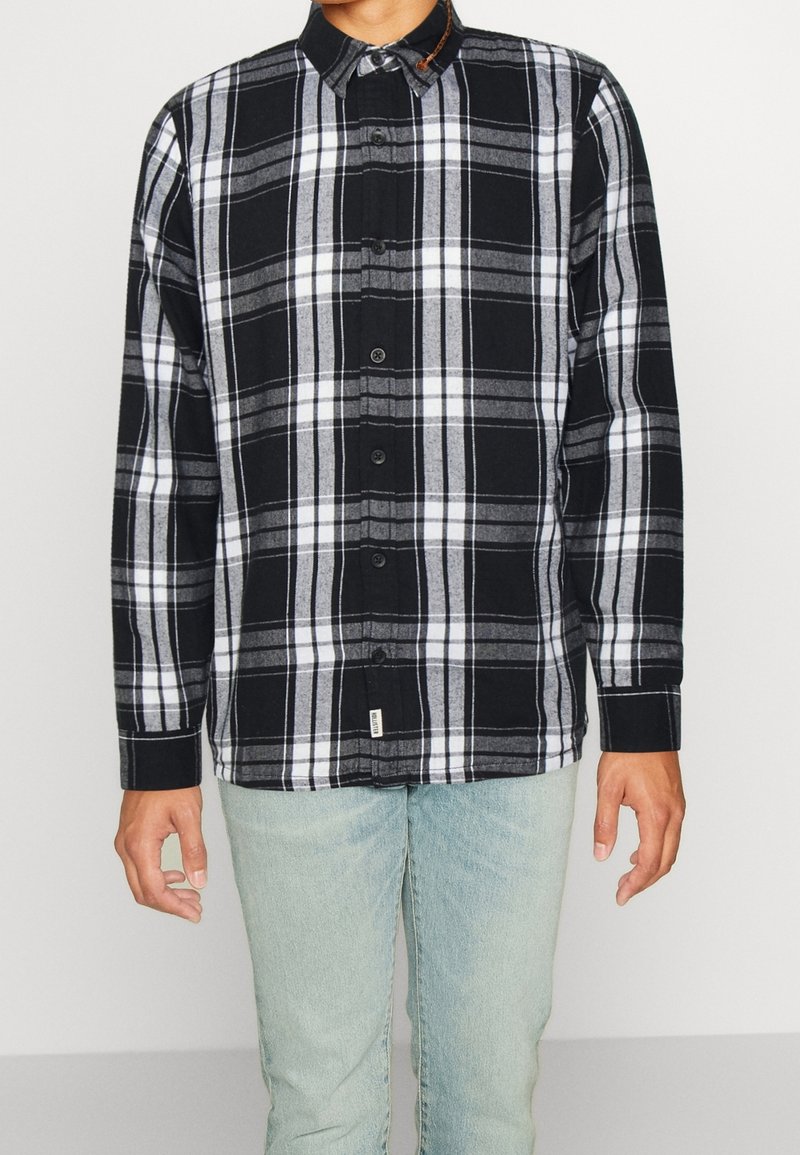 Svart och vit rutig skjorta med grå inslag, knappad krage, lång ärm och en sydd etikett nära fållen, matchad med ljusblå jeans.