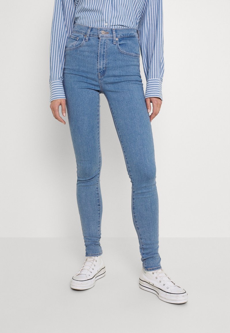 Personne portant un jean skinny taille haute bleu clair, une chemise rayée bleue et blanche à manches longues, et des baskets blanches montantes, debout devant un fond uni.