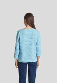 Helles blaues Oberteil aus strukturiertem Stoff mit horizontalem Rüschendesign, verfügt über Dreiviertelärmel und einen runden Ausschnitt. Kombiniert mit dunklen Jeans.