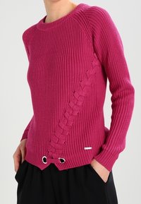 Frau, die einen leuchtend magentafarbenen Strickpullover mit einer diagonalen geflochtenen Detailierung und Ösen am Saum trägt, kombiniert mit schwarzen Hosen.