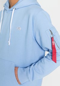 Felpa lilla chiaro realizzata in morbido tessuto. Presenta una tasca frontale, lacci bianchi e una tasca con zip sulla manica con un accento di nastro rosso.