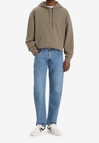 Olivgrön hoodie med känguruficka och dragsko, kombinerad med ljusblå raka jeans och vita sneakers med svarta accenter.