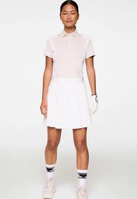 Jasnobeżowa koszulka polo z białymi bocznymi panelami, zestawiona z białą plisowaną spódnicą. Dodatki to biała rękawica golfowa i jasne tenisówki.