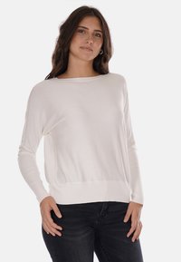 Maglione bianco a maniche lunghe, con taglio morbido e collo a barca, bordo a costine. Tessuto con texture, abbinato a jeans neri.