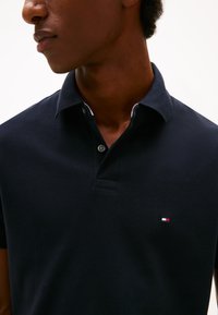 Polo azul-marinho com tecido texturizado, gola com botões e pequeno detalhe de logótipo no peito. Mangas curtas e design clássico.