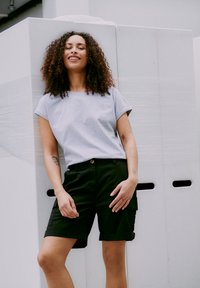 Kvinna med lockigt hår iförd en ljusgrå t-shirt och svarta shorts, stående och leende mot en hög av stora vita lådor.