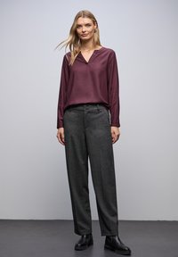 Burgunderfarbene Bluse mit langen Ärmeln und V-Ausschnitt, kombiniert mit anthrazitfarbenen weiten Hosen und schwarzen Stiefeletten, vor einem hellgrauen Hintergrund.