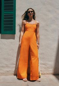 Une combinaison orange avec un corsage ajusté, des bretelles spaghetti et des broderies florales. Conception à jambes larges en tissu léger, associée à des sandales plates.