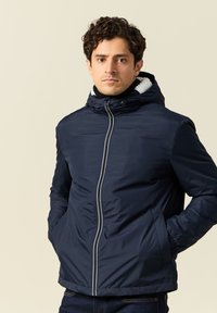 Giacca imbottita blu navy con cappuccio, dotata di zip anteriore, polsini elasticizzati e tasche laterali. Foderata con un morbido materiale bianco all'interno.