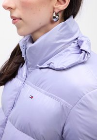 Chaqueta acolchada de color púrpura claro con cuello alto y capucha. Presenta un pequeño logo bordado en el pecho y una textura lisa y brillante.