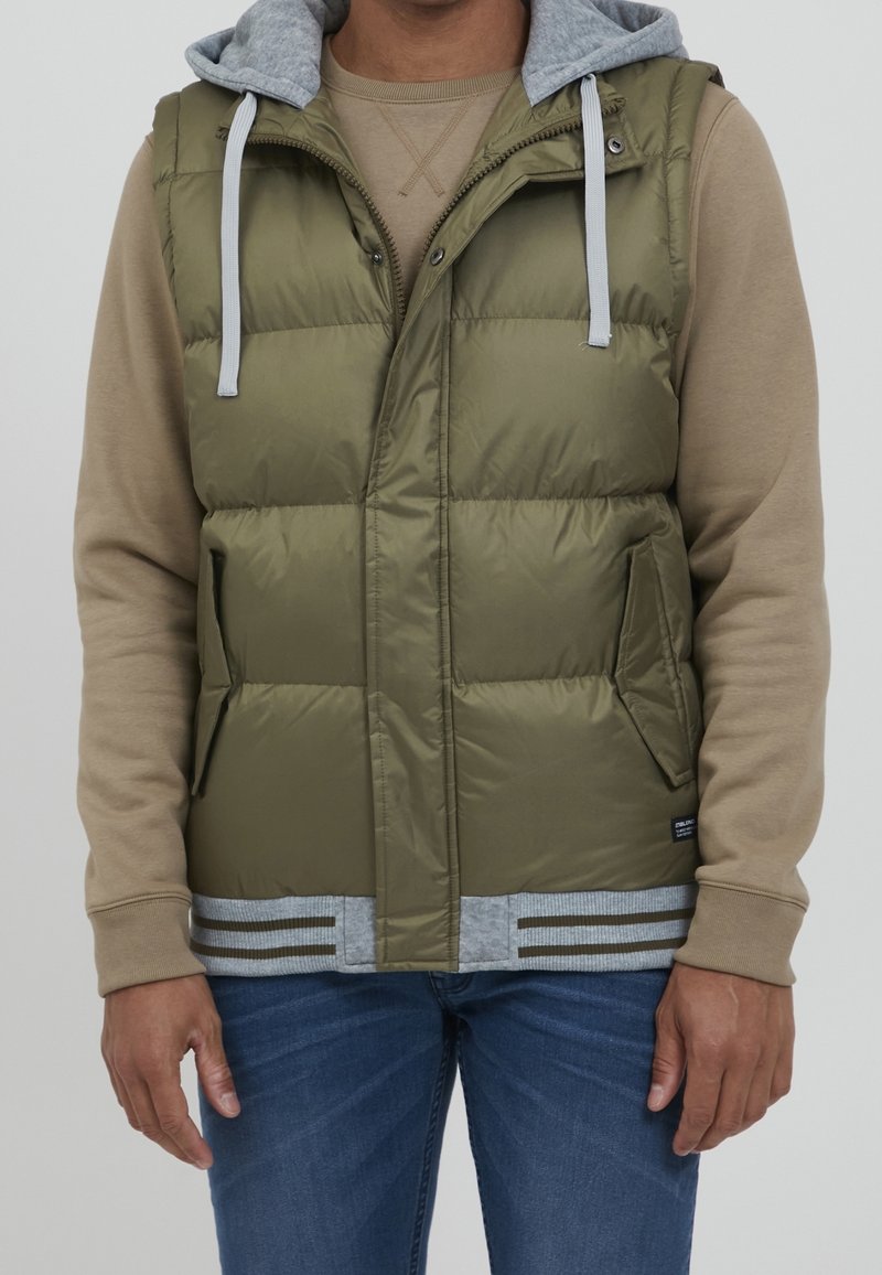 Homme portant une veste matelassée sans manches vert olive avec une capuche grise, sur une chemise à manches longues beige et un jean bleu, debout les bras détendus.