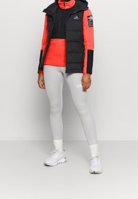 Gilet nero imbottito con cappuccio sopra una maglietta a maniche lunghe rossa brillante. Leggings grigio chiaro e sneakers bianche con dettagli testurizzati.