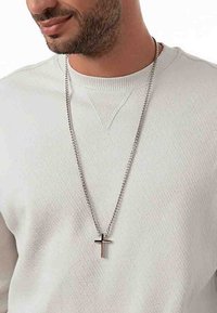 Silver kedja halsband med ett kors hänge, med kontrasterande guldfärgade accenter. Bärs över en ljusfärgad, texturerad tröja.