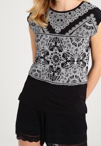 Schwarzes T-Shirt mit komplizierten, weißen Spitzenmustern im oberen Bereich und einem einfarbig schwarzen unteren Abschnitt. Kurze Ärmel und runder Ausschnitt.