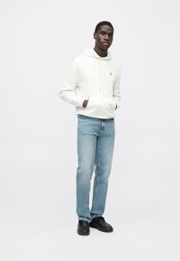 Sudadera blanca con capucha y bolsillo frontal, con un pequeño logo rojo, combinada con vaqueros azul claro y calzado negro. Diseño sencillo y casual.