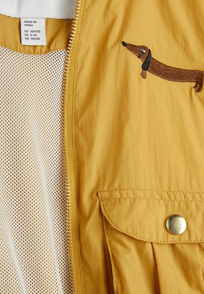 Rumena jakna z zadrgo, lahka tkanina, mrežasta podloga in vezenina rjavega taksija na sprednji strani.