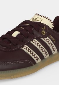 Zbliżenie ciemnobrązowego buta sportowego Adidas Samba z kremowymi paskami, przeszyciami, gumową podeszwą i złotymi przelotkami.
