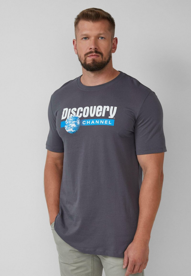 Grijze katoenen t-shirt met het "Discovery CHANNEL" logo in wit en blauw, met een cirkelvormige aarde-afbeelding op de voorkant. Korte mouwen, ronde hals.