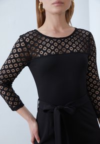 Vestido negro con mangas largas de encaje que presentan un patrón geométrico, cintura ceñida con un cinturón de lazo y un cuerpo de tela suave.