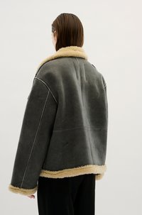 Grijze schapenvachtjas met een tan nepbont kraag en bies. Kenmerkt zich door een losse pasvorm en zichtbare naaddetails op de rug.