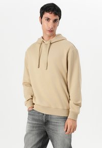 Camisola bege com cordão, textura suave, punhos e barra canelados. Combinada com jeans cinzentos desbotados, de pé contra um fundo simples.