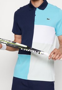 Koszulka polo w granatowe, jasnoniebieskie, białe i turkusowe kolory w blokach. Posiada kołnierzyk, guziki i małe logo. Trzyma rakietę tenisową.