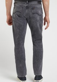 Man som bär svarta tvättade jeans med två bakfickor och kors tatueringar på båda underarmarna, står mot en vit bakgrund.