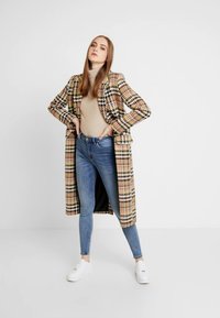 Μακριά καρό καπαρτίνα σε μπεζ, μαύρους και κόκκινους τόνους, φορεμένη πάνω από μπεζ ζιβάγκο, συνδυασμένη με εφαρμοστά μπλε τζιν και λευκά αθλητικά.