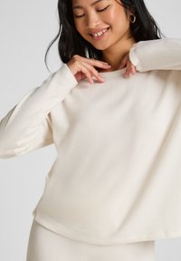 Witte geribde top met lange mouwen en een relaxte pasvorm, voorzien van een bootnek en een zachte textuur. Model toont de drape en beweging van de top.