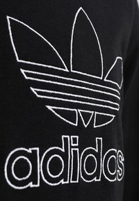 Černá bavlněná látka s velkým bílým vyšívaným logem Adidas ve tvaru trojlístku se třemi listy a vodorovnými pruhy nad názvem značky.