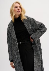 Čierno-biely kabát v károvanom vzore z texturovaného materiálu, s voľným strihom, širokými klopami a bočnými vreckami. Kombinovaný s rolákom.