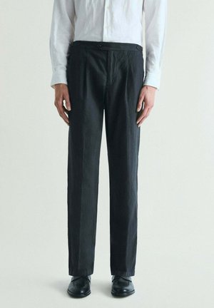 Hombre con pantalones negros con botones en la cintura, camisa blanca de manga larga y zapatos negros, de pie contra un fondo claro y sencillo.