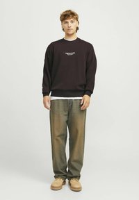 Zwarte sweatshirt met wit logo, losse olijfgroene jeans en beige schoenen. De outfit heeft eenvoudige ontwerpen en neutrale kleuren.