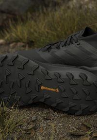 Chaussures de randonnée noires sur un sol rocailleux montrant la semelle extérieure robuste Continental avec un logo orange dans un environnement extérieur avec de l'herbe et des feuilles.