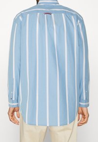 Camisa azul claro de manga larga con rayas blancas verticales, puño abotonado y dobladillo redondeado. Incluye una marca sutil en la manga.