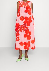 Vestido midi sin mangas de tela rosa con grandes estampados florales rojos. Combinado con zapatos de tacón verde de punta, mostrando un look vibrante.