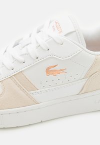 Fehér bőr sportcipő bézs velúr részletekkel, perforált díszítéssel és a domború korall Lacoste logóval. Fehér fűzőkkel és gumitalppal rendelkezik.