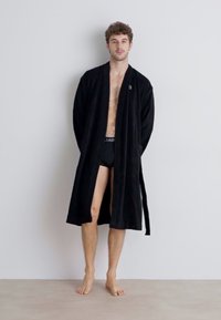 Robe preto até aos joelhos, com um tecido texturizado, apresentando um fecho de laço na cintura e dois bolsos laterais. Usado sobre boxers pretos.