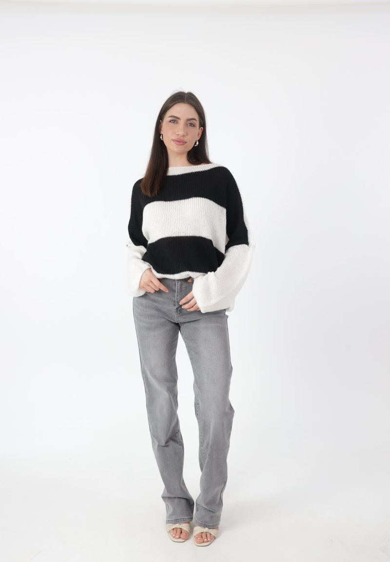 Maglione a righe bianche e nere in maglia con maniche ampie, abbinato a jeans dritti grigio chiaro e sandali nudi su uno sfondo neutro.