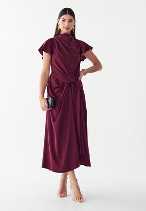 Donna che indossa un vestito midi color burgundy con maniche corte svolazzanti, orecchini dorati, braccialetto, tiene una pochette nera e indossa tacchi dorati con cinturino alla caviglia.