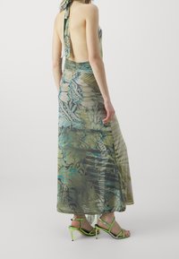 Maxiklänning med blommönster i gröna och blå nyanser, halterneckdesign, rygglös stil, matchad med gröna sandaletter med hög klack och remmar.