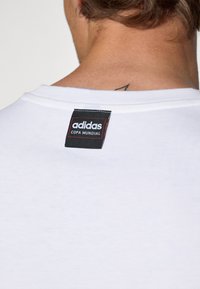 Hvid bomulds-t-shirt med et sort mærke med hvid og rød tekst, der læser 'adidas COPA MUNDIAL', placeret bag på nakken.