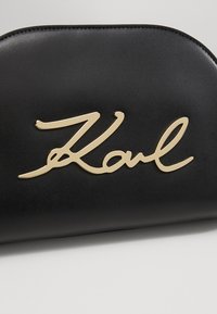 Czarna torba z ekoskóry z logo „Karl” w złotym kursywie na przodzie. Zaokrąglony kształt, gładka faktura i przeszycia wzdłuż krawędzi.