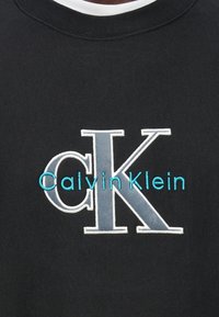 Μαύρο φούτερ με έντονο κεντημένο λογότυπο. Το λογότυπο αποτελείται από ένα μεγάλο «CK» σε γκρι και τιρκουάζ, με το «Calvin Klein» από κάτω.