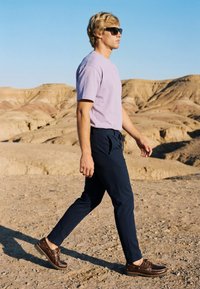 Jeune homme aux cheveux blonds et lunettes de soleil marchant sur un terrain désertique rocheux, portant un t-shirt violet clair, un pantalon foncé et des chaussures en cuir marron.
