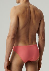 Męskie kąpielówki w kolorze koralowym; gładki, elastyczny materiał; niskie talii; minimalne zakrycie z bezszwowym pasem.