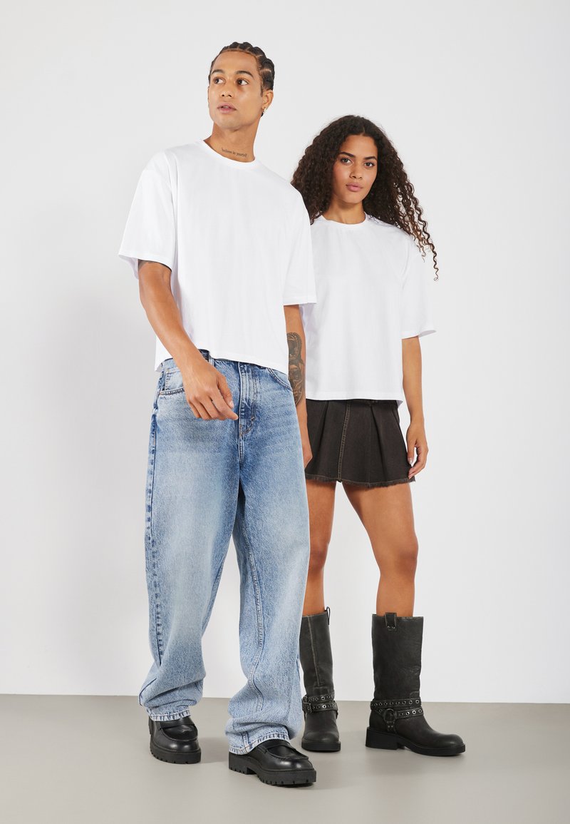 To modeller iført hvite, oversized t-skjorter. En i lys blå vide jeans, den andre i et svart plisserte skjørt og høye svarte støvler.