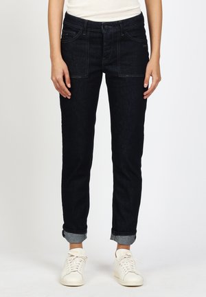 Person trägt schwarze umgeschlagene Jeans und weiße Turnschuhe, steht mit entspannten Händen an den Seiten vor einem einfarbigen Hintergrund.